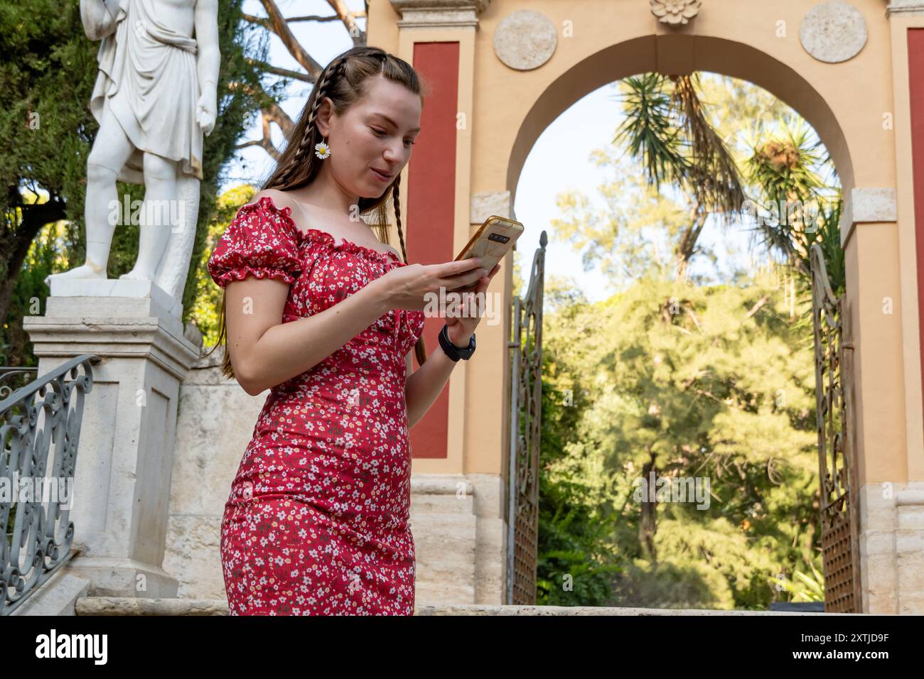 Eine junge rothaarige Frau in einem roten Blumenkleid schickt eine Textnachricht auf ihrem Smartphone, während sie einen sonnigen Tag im Freien genießt Stockfoto
