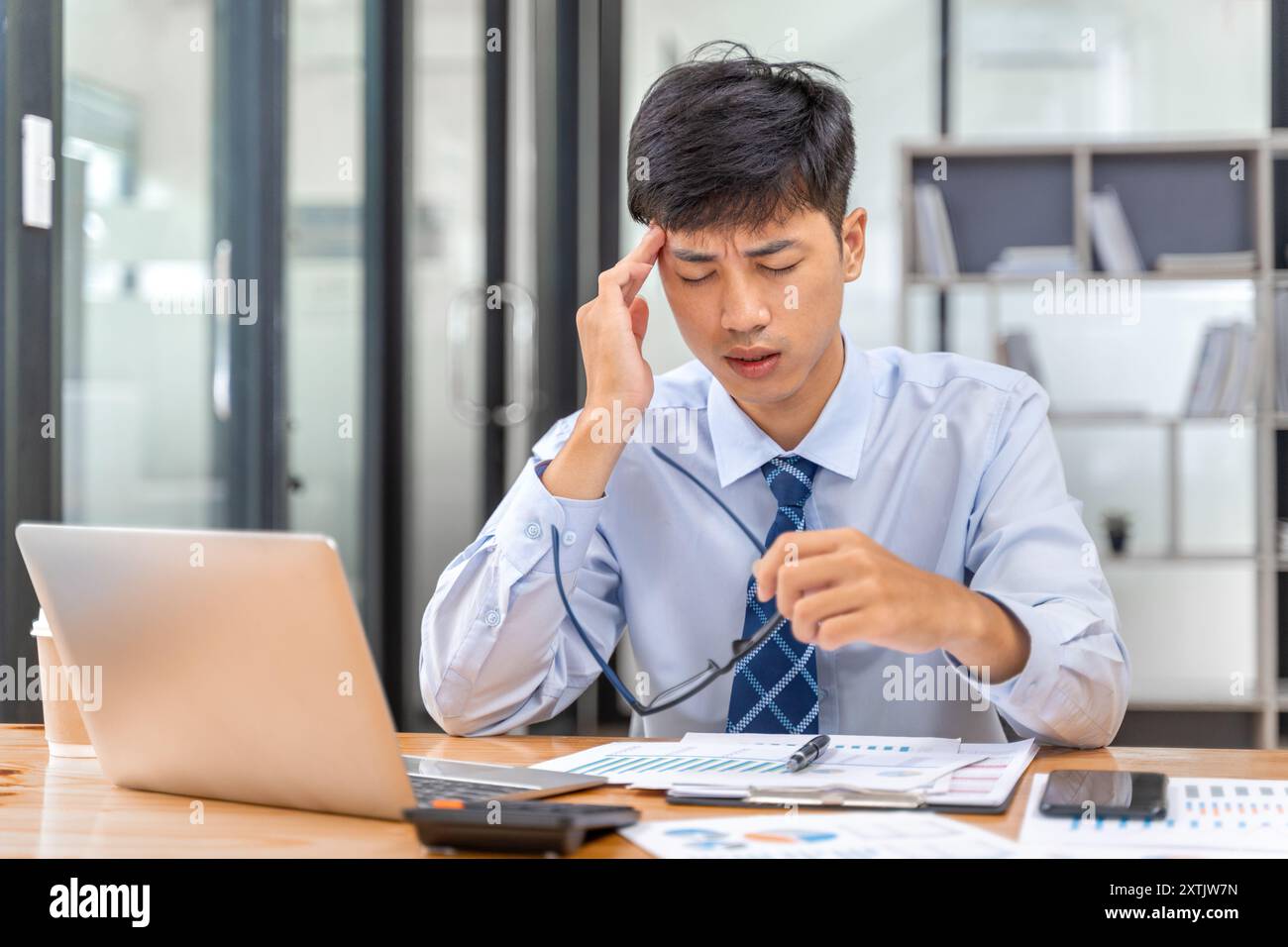 Erschöpfter junger Geschäftsmann, der in seinem Büro sitzt und seine Hand auf den Kopf legt, um die Schmerzen seiner intensiven Kopfschmerzen zu lindern Stockfoto