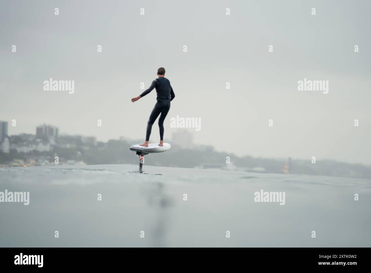 Ein junger Mann in einem Neoprenanzug reitet im Meer und fliegt über dem Wasser auf einem elektrisch betriebenen Tragflächenbrett, einer Hybridfolie mit Eoil-Folienantrieb Stockfoto