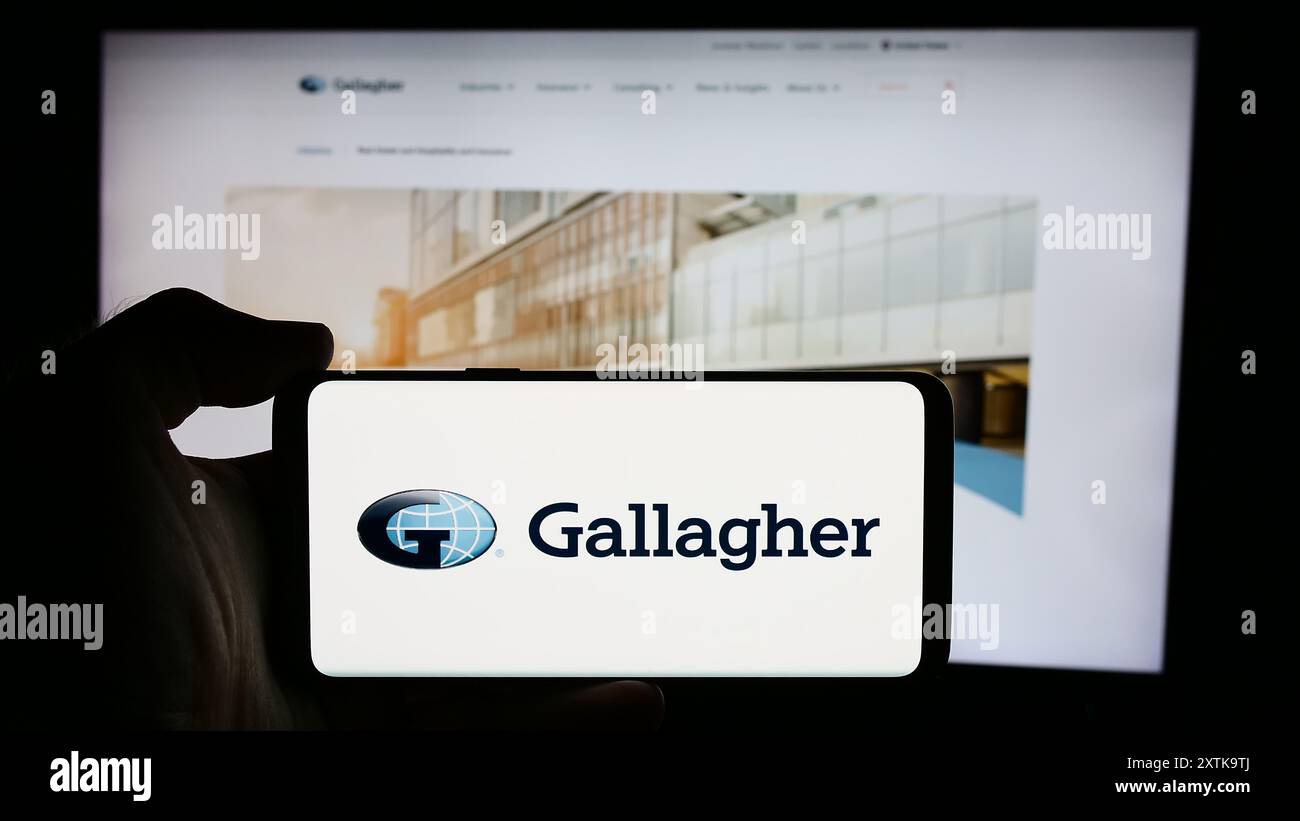 Person, die ein Mobiltelefon mit dem Logo des US-Versicherungsunternehmens Arthur J. Gallagher Co. (AJG) vor der Website des Unternehmens hält. Schwerpunkt auf dem Display des Telefons. Stockfoto