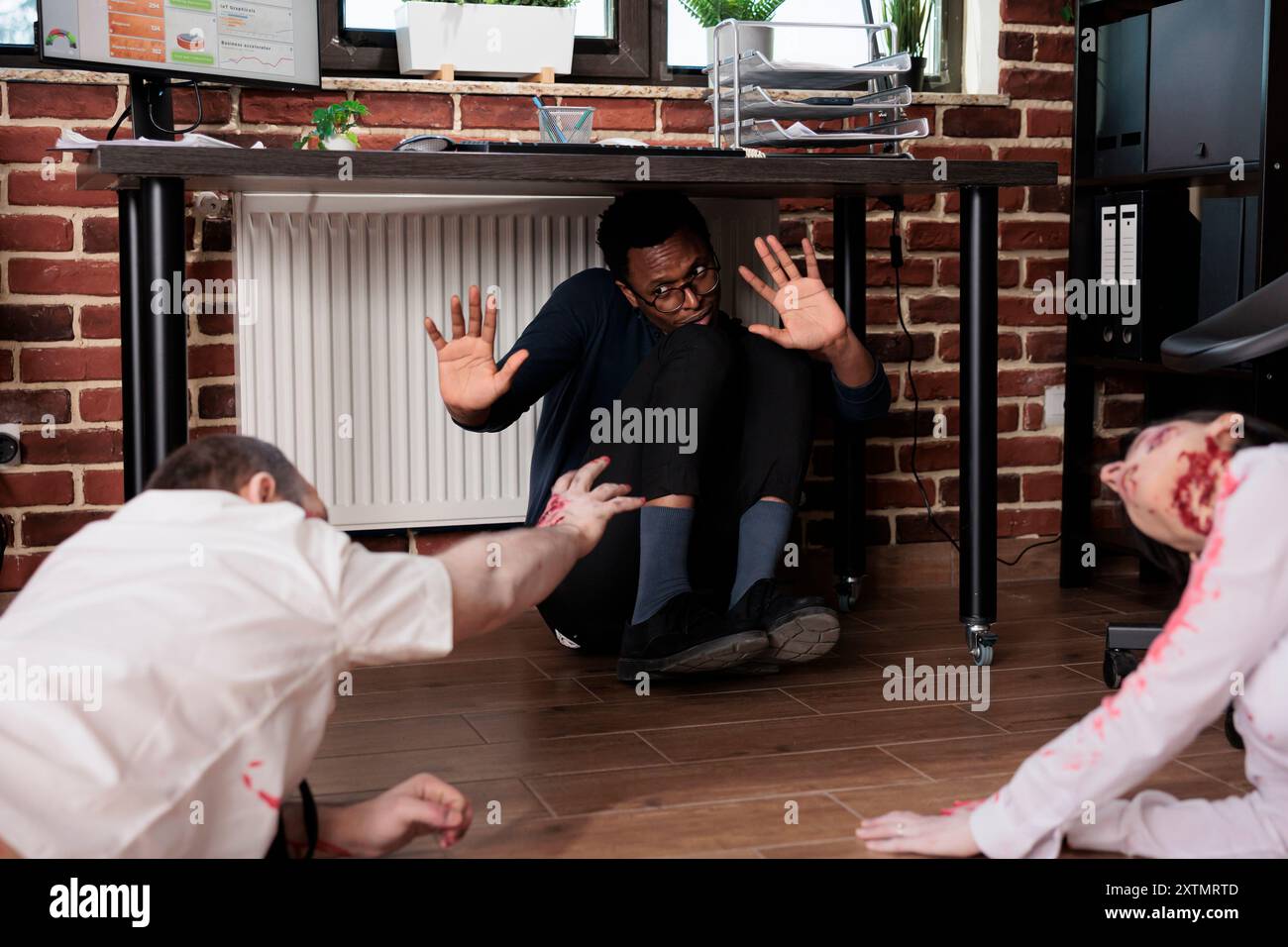 Schauspieler, die Zombies in der Horrorfilmszene spielen, schnappen sich BIPOC-Arbeiter, der sich unter dem Schreibtisch im Büroset versteckt. Mann und Frau mit reanimierten Leichen Rollen in Thriller-Film Attacking Opfers Stockfoto