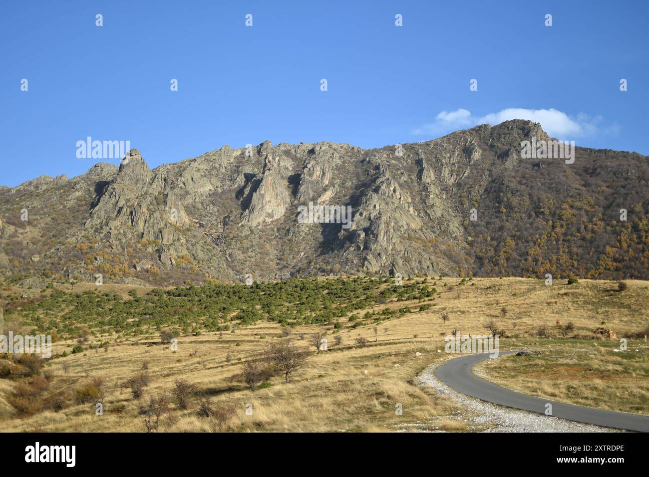 Majestätischer felsiger Berg unter klarem blauem Himmel, eine gewundene Straße durchquert das zerklüftete Gelände und unterstreicht die ruhige Schönheit der Natur. Stockfoto