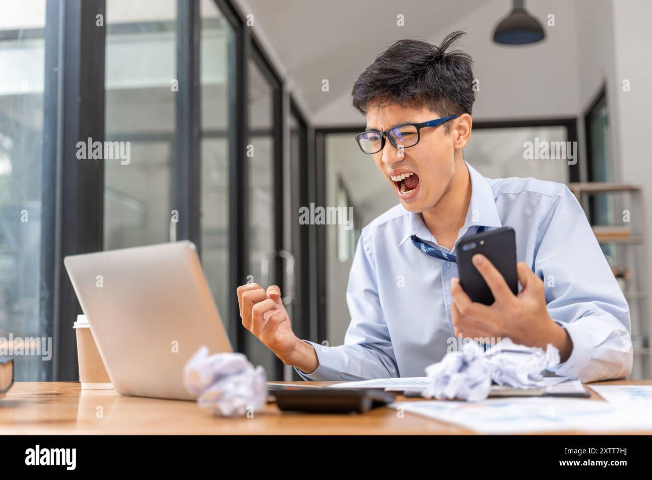 Frustrierter und wütender junger Geschäftsmann, der spät im Büro arbeitet und versucht, sein Projekt zu beenden, indem er ein Telefon in einer Hand hält und wütend auf den Bildschirm schaut Stockfoto