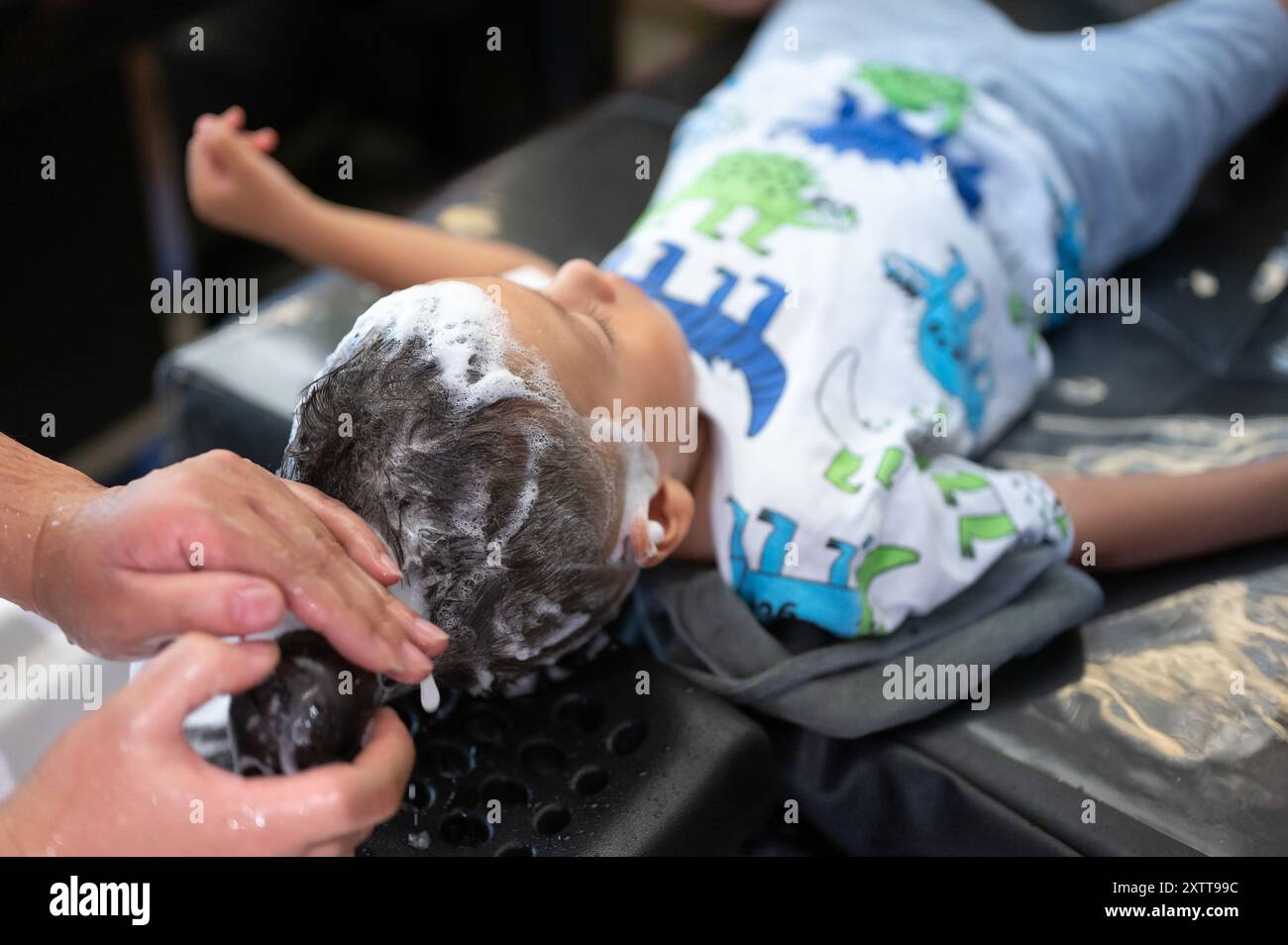 Auf diesem Bild legt sich ein Kind hin, während seine Haare in einem Friseursalon gewaschen werden. Das Kind trägt ein farbenfrohes Outfit mit Dinosauriermotiv. Der Schwerpunkt liegt auf Stockfoto