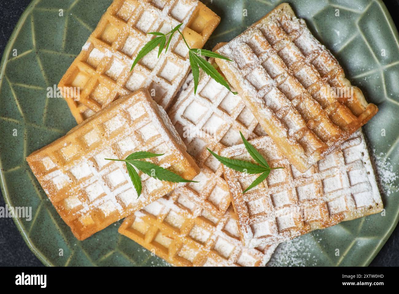 Köstliche Waffeln mit Puderzucker bestreut und mit frischen Marihuanablättern auf einem grünen Teller dekoriert. Nahaufnahme, Draufsicht Stockfoto