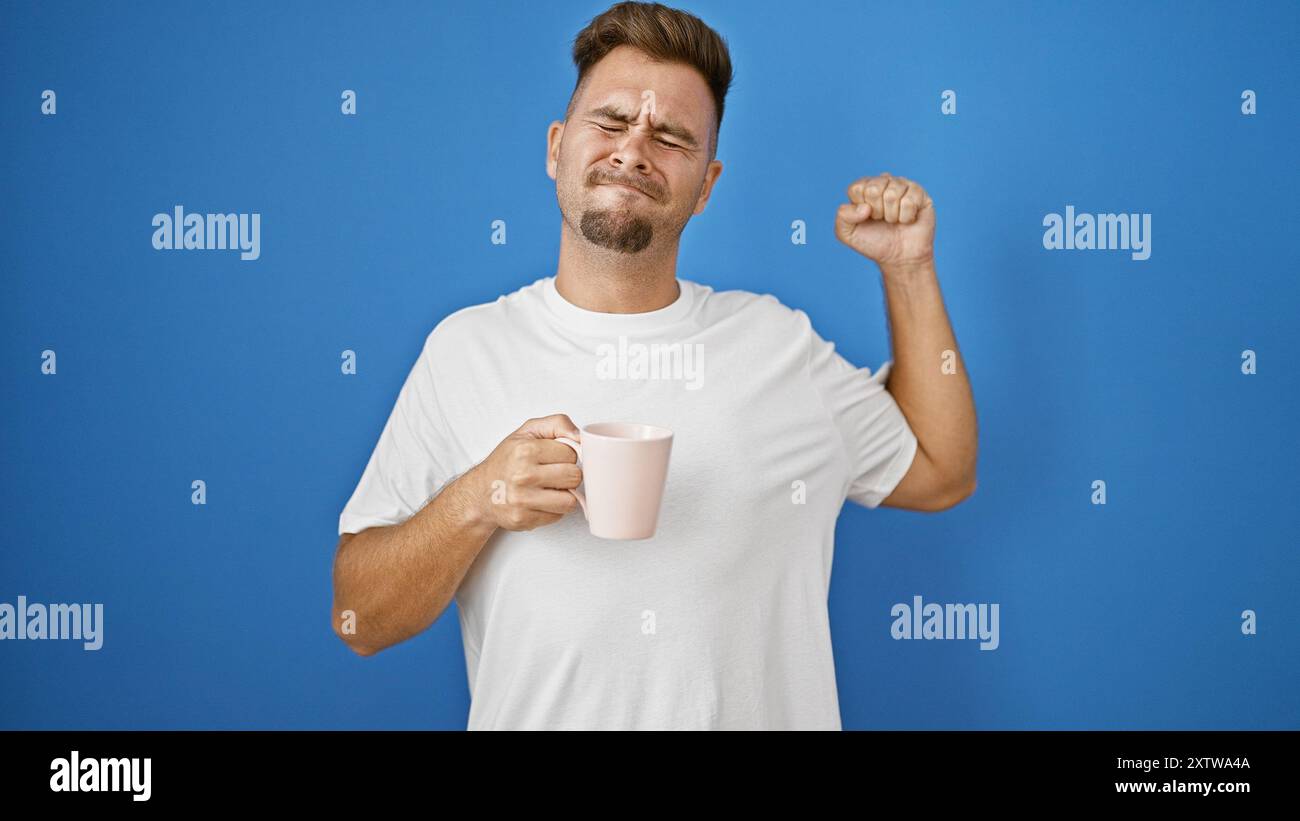 Ein junger hispanischer Mann dehnt sich mit Unwohlsein an einer blauen Wand aus und hält einen rosa Becher. Stockfoto