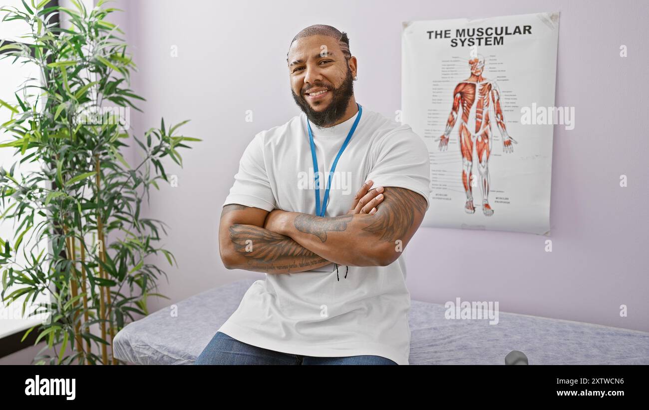 Lächelnder bärtiger Mann mit Tattoos und Schlüsselband, der in einer Klinik steht, mit einem muskulären System-Poster im Hintergrund. Stockfoto