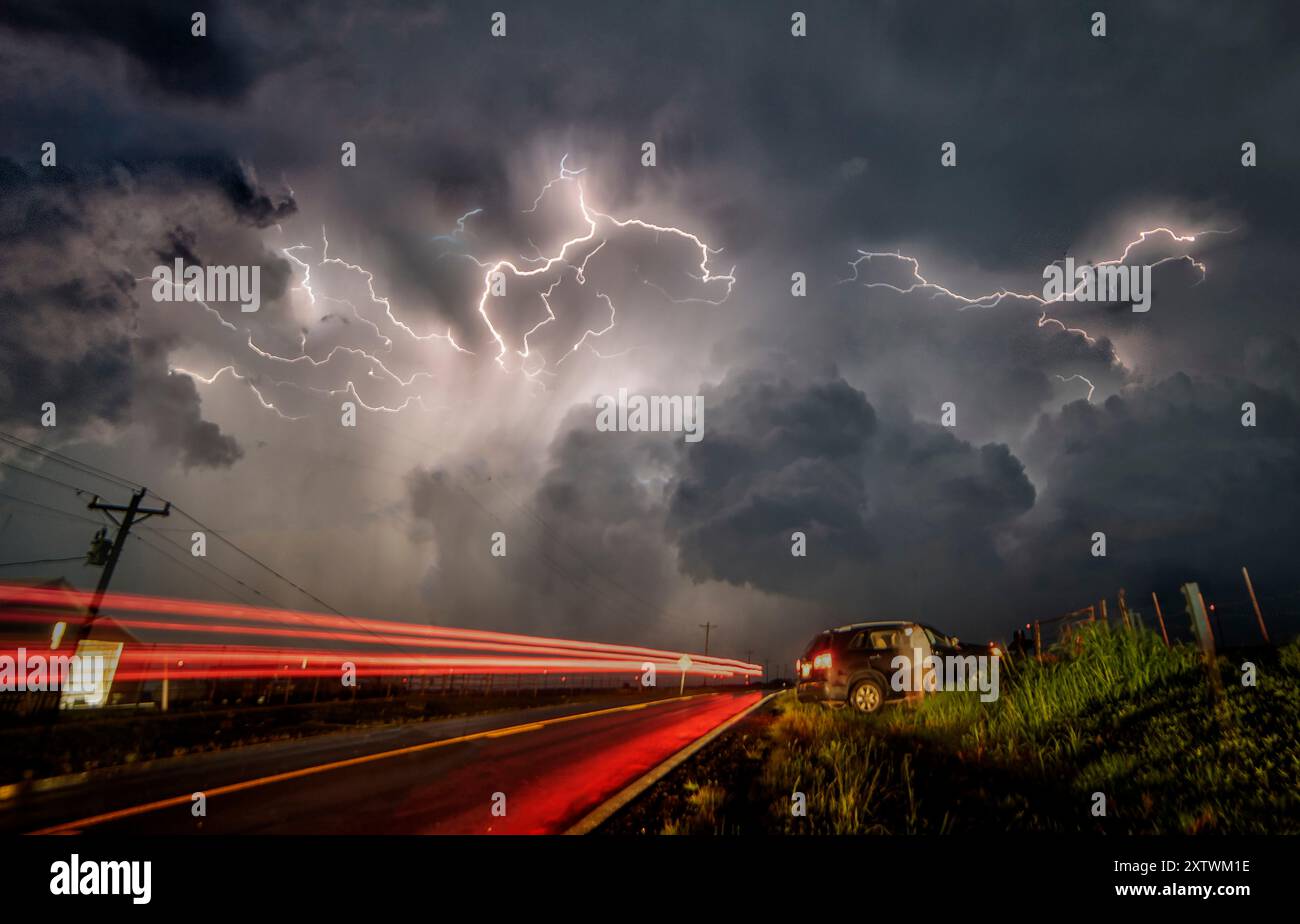 Ein intensiver Gewitter beleuchtet den Himmel über einer Straße, wenn Fahrzeuge vorbeifahren, und erzeugt leichte Wege in einer dramatischen und turbulenten Atmosphäre. Stockfoto