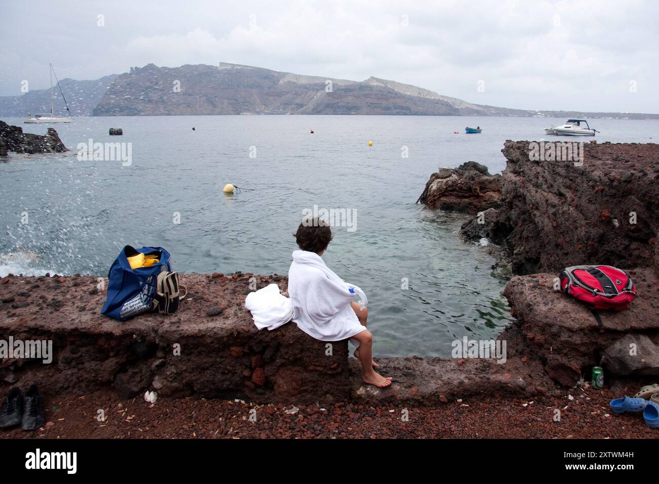 Eine Person, die in ein weißes Handtuch gewickelt ist, sitzt auf einem felsigen Felsvorsprung am Meer, mit Blick auf eine malerische Aussicht mit Booten und Bergen im Hintergrund. Stockfoto