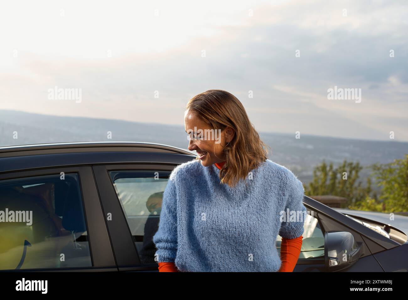 Eine lächelnde Frau in einem blauen Pullover steht neben einem Auto mit einem malerischen Ausblick im Hintergrund. Stockfoto