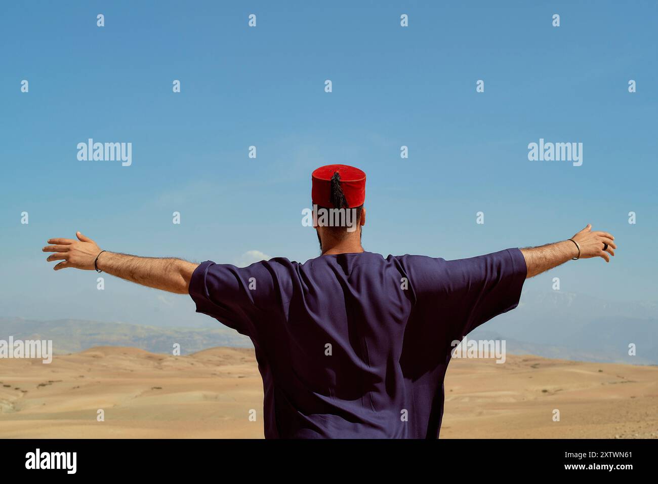 Ein Mann in traditioneller Kleidung mit offenen Armen, der vor einer riesigen Wüstenlandschaft unter einem klaren blauen Himmel steht. Stockfoto