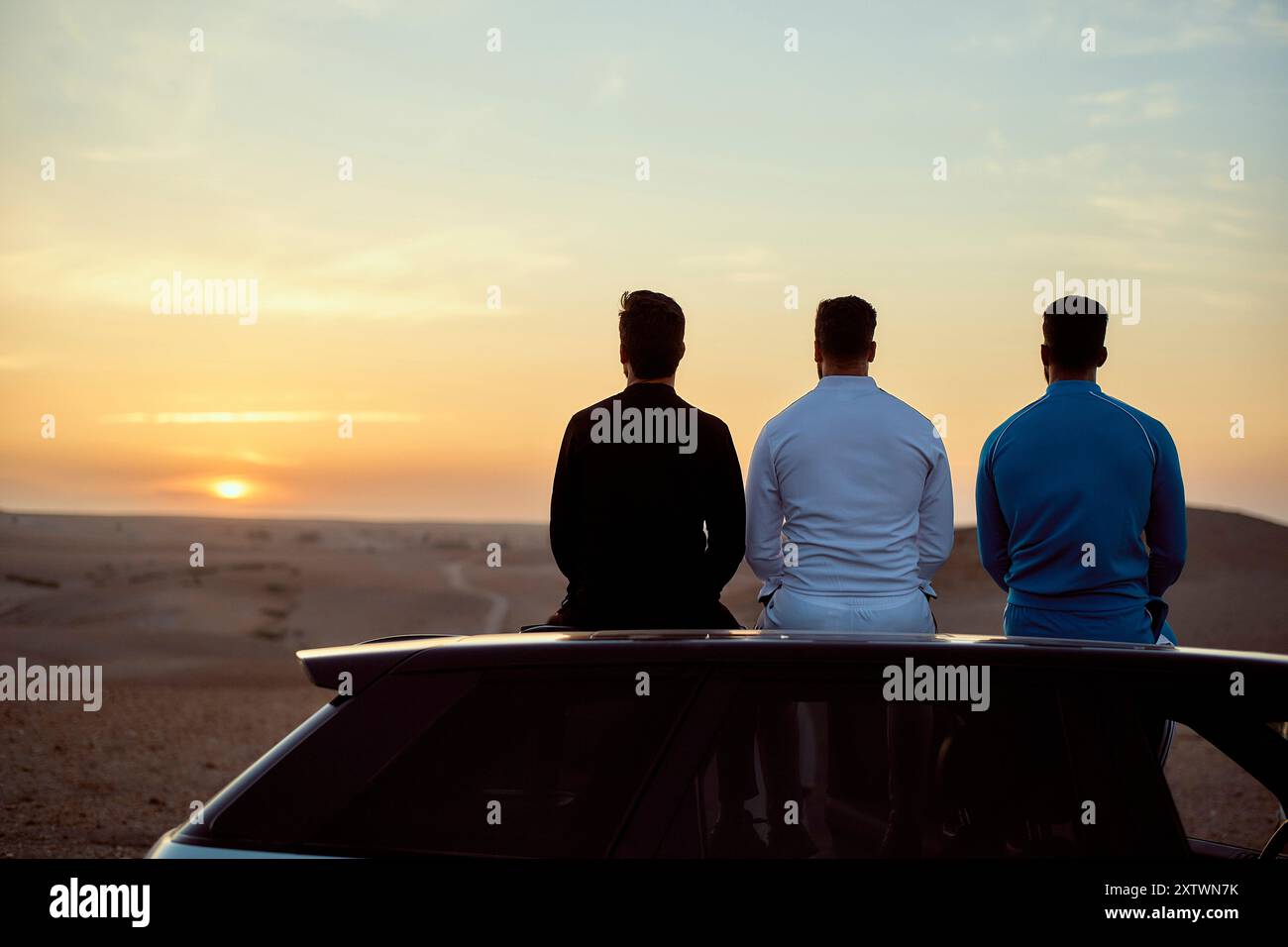 Drei Männer sitzen auf einem Auto und beobachten den Sonnenuntergang in der Wüste. Stockfoto