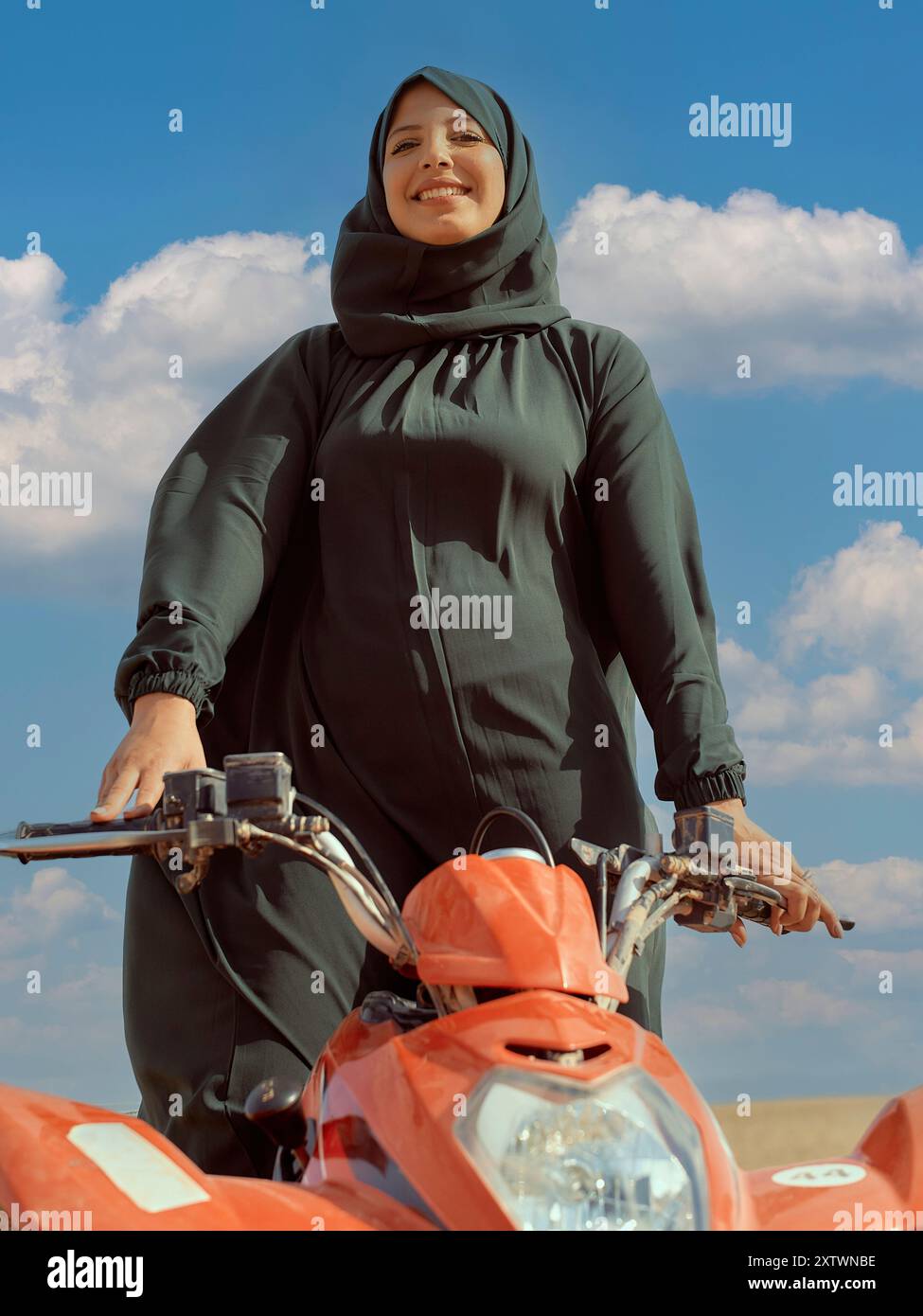 Lächelnde Frau in einem Hijab, die mit den Händen am Lenker eines roten Motorrads unter einem klaren blauen Himmel steht. Stockfoto