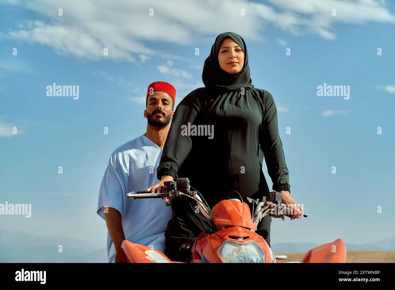 Eine Frau in einem Hijab, die selbstbewusst hinter einem Mann auf einem roten Motorrad vor einem klaren Himmel steht. Stockfoto