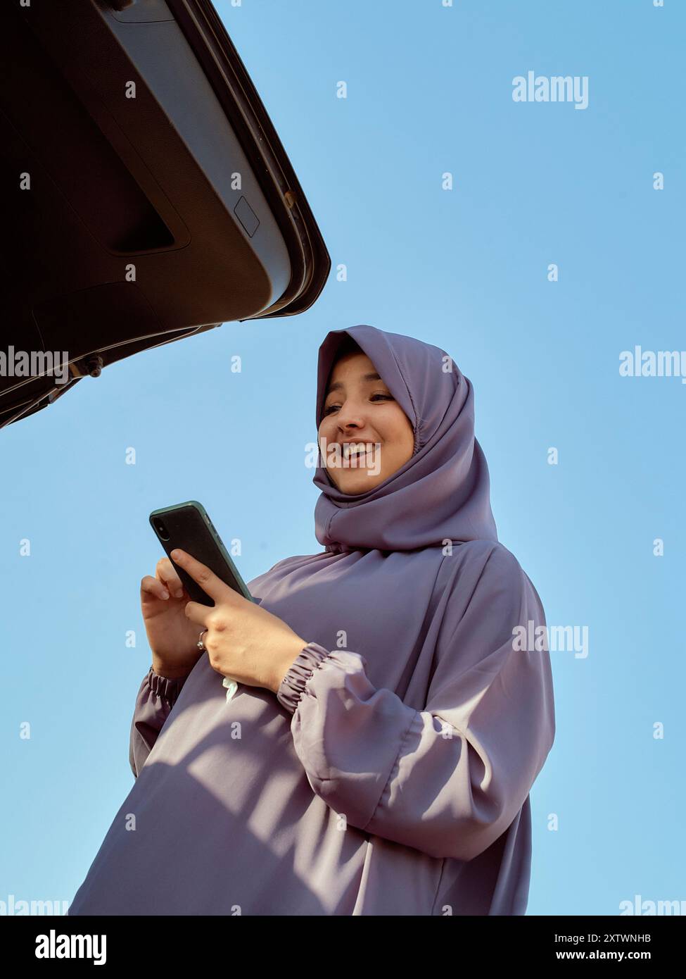 Lächelnde Frau in einem Hijab, die ein Smartphone mit geöffnetem Kofferraum im Hintergrund benutzt. Stockfoto