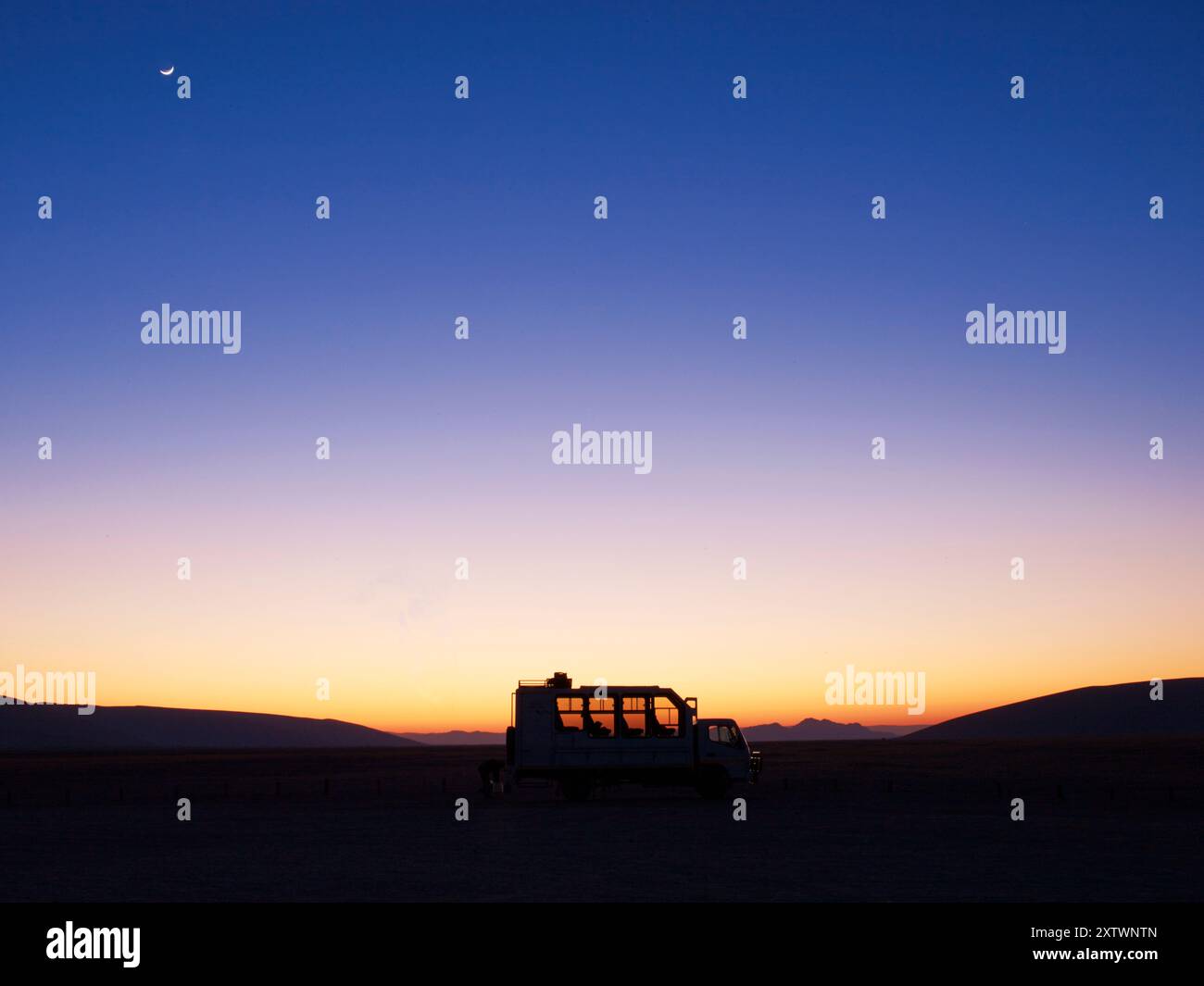 Ein ruhiger Himmel in der Abenddämmerung mit Gradienten von Orange und Violett über der Silhouette eines Wohnwagens in einer weiten, offenen Landschaft unter einem Halbmond. Stockfoto