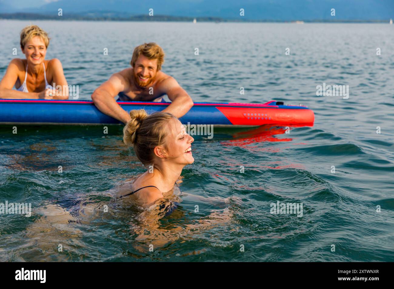 Drei Freunde genießen einen Tag am See mit einer Frau im Vordergrund und zwei Personen auf einem Paddleboard im Hintergrund. Stockfoto