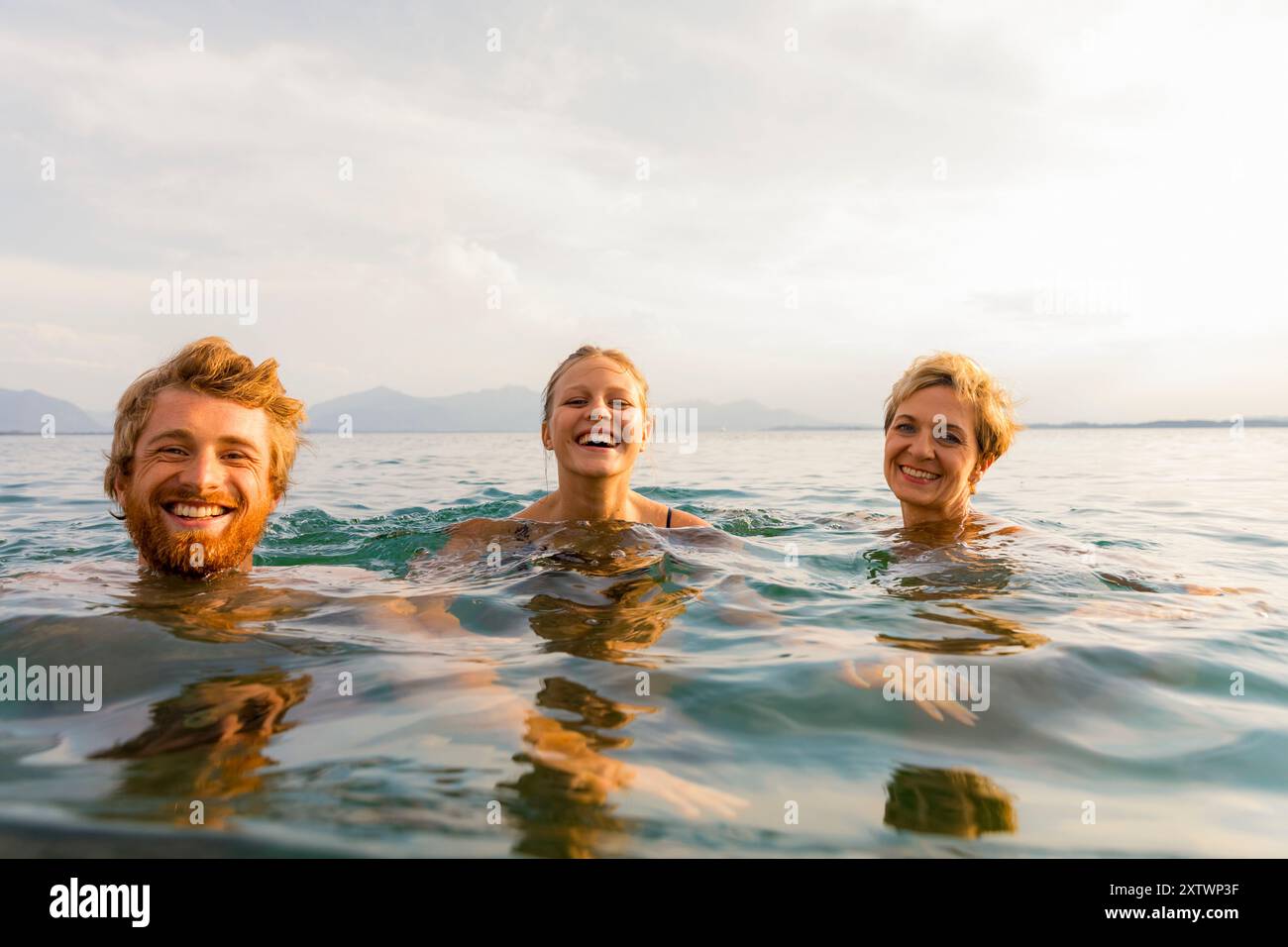 Drei Personen lächeln und schwimmen im offenen Wasser mit Bergen im Hintergrund unter klarem Himmel. Stockfoto