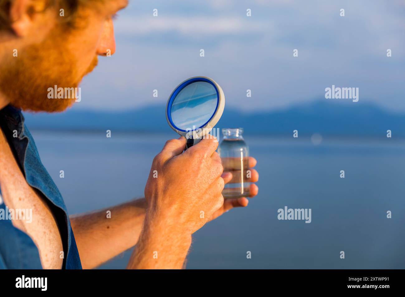 Mann untersucht Wasser in einer kleinen, transparenten Flasche mit einer Lupe vor einer ruhigen Seeblicke. Stockfoto