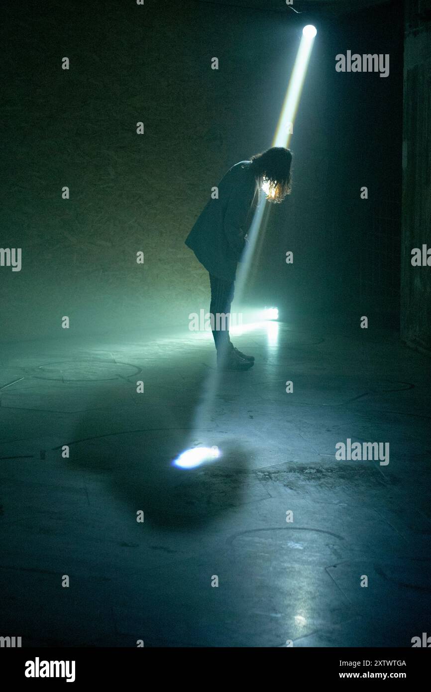 Ein einsamer Mensch steht gebadet in einem Lichtstrahl, mit gebeugtem Kopf, in einem verdunkelten Raum. Stockfoto