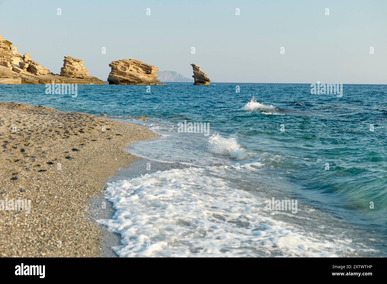 Ein ruhiger Strand mit klarem, blauem Wasser, das sanft gegen eine Sandküste schlägt, unter einem klaren Himmel mit zerklüfteten Felsformationen in der Ferne. Stockfoto