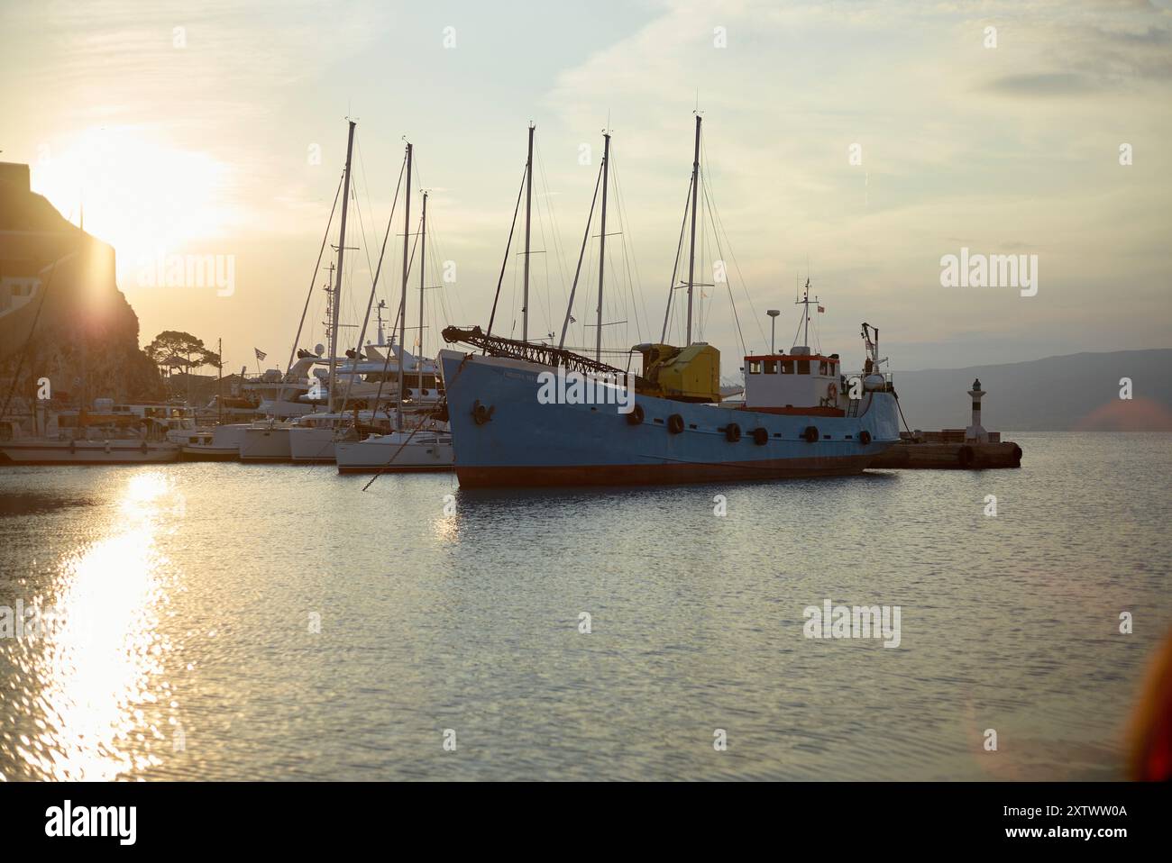 Blick am frühen Morgen auf einen ruhigen Hafen mit einem blauen Boot, das neben einem Pier liegt, mit sanftem Sonnenlicht, das vom ruhigen Wasser glitzert. Stockfoto