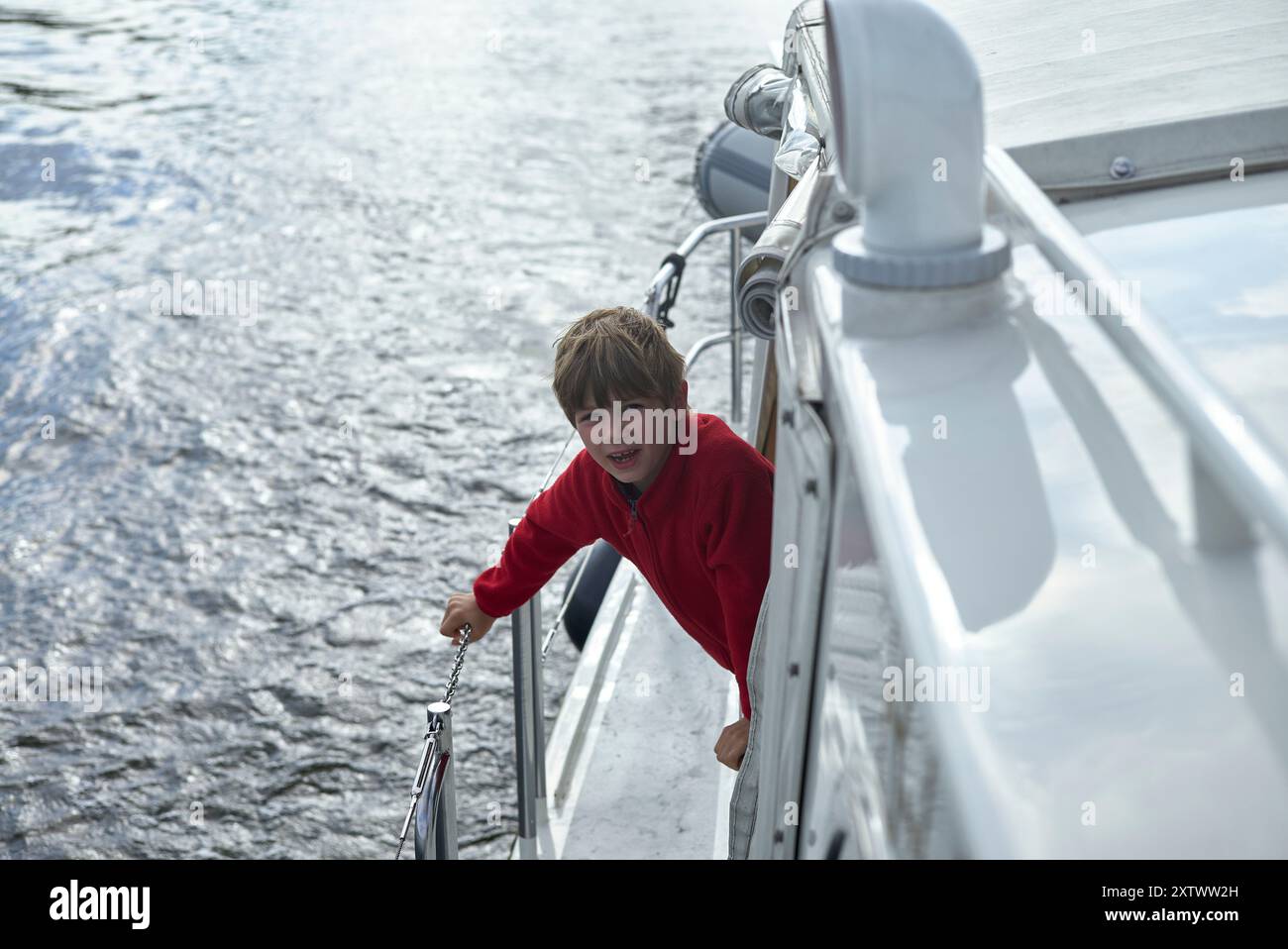 Kind in roter Jacke, das an der Seite eines Bootes mit Wasser im Hintergrund hält. Stockfoto