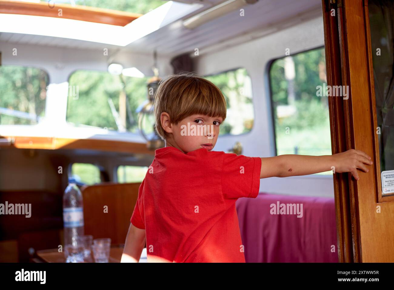 Junge in einem roten Hemd, der in einem hellen Wohnmobil steht und sich umdreht, um die Kamera mit einem nachdenklichen Ausdruck zu betrachten. Stockfoto