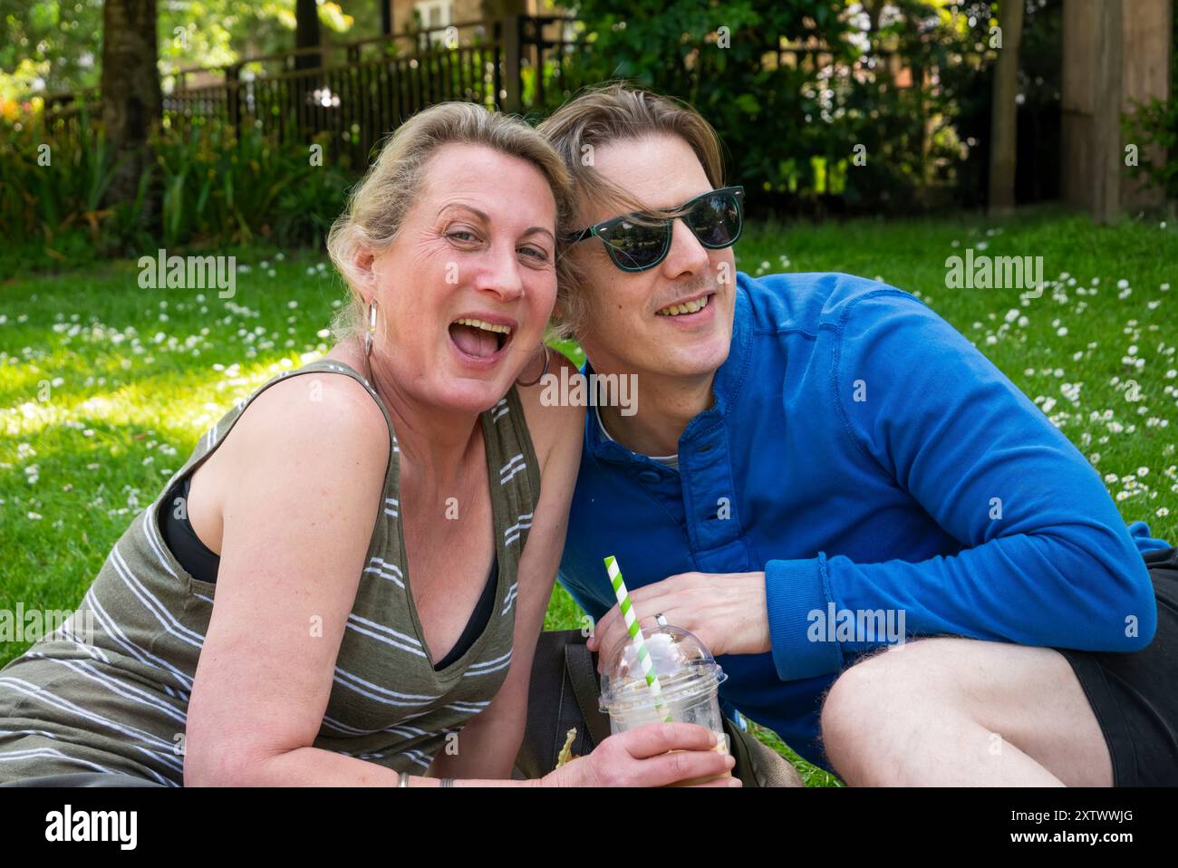 Fröhliche Frau und Mann sitzen auf dem Gras in einem Park mit Gänseblümchen und genießen einen sonnigen Tag mit erfrischenden Getränken. Stockfoto