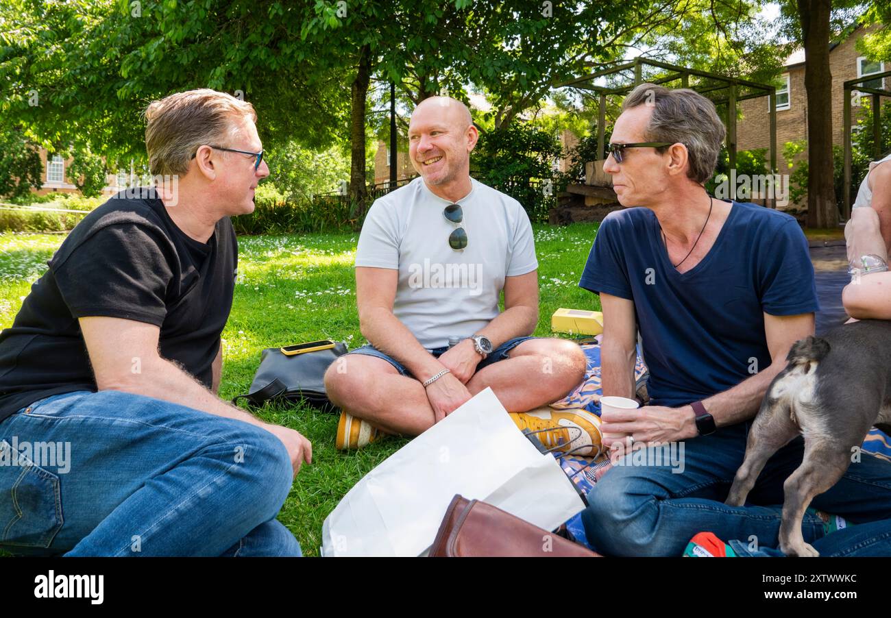 Drei Männer und ein kleiner Hund unterhalten sich an einem sonnigen Tag im Park, sitzen mit sichtbaren Picknickstücken auf dem Gras. Stockfoto