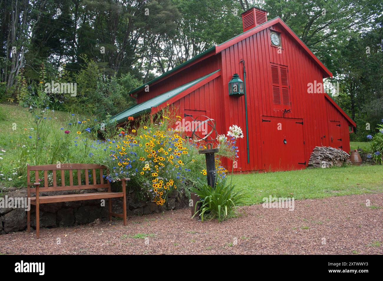 Eine lebendige rote Scheune, umgeben von üppigem Grün und bunten Blumen mit einer Holzbank im Vordergrund, Auckland, Neuseeland Stockfoto