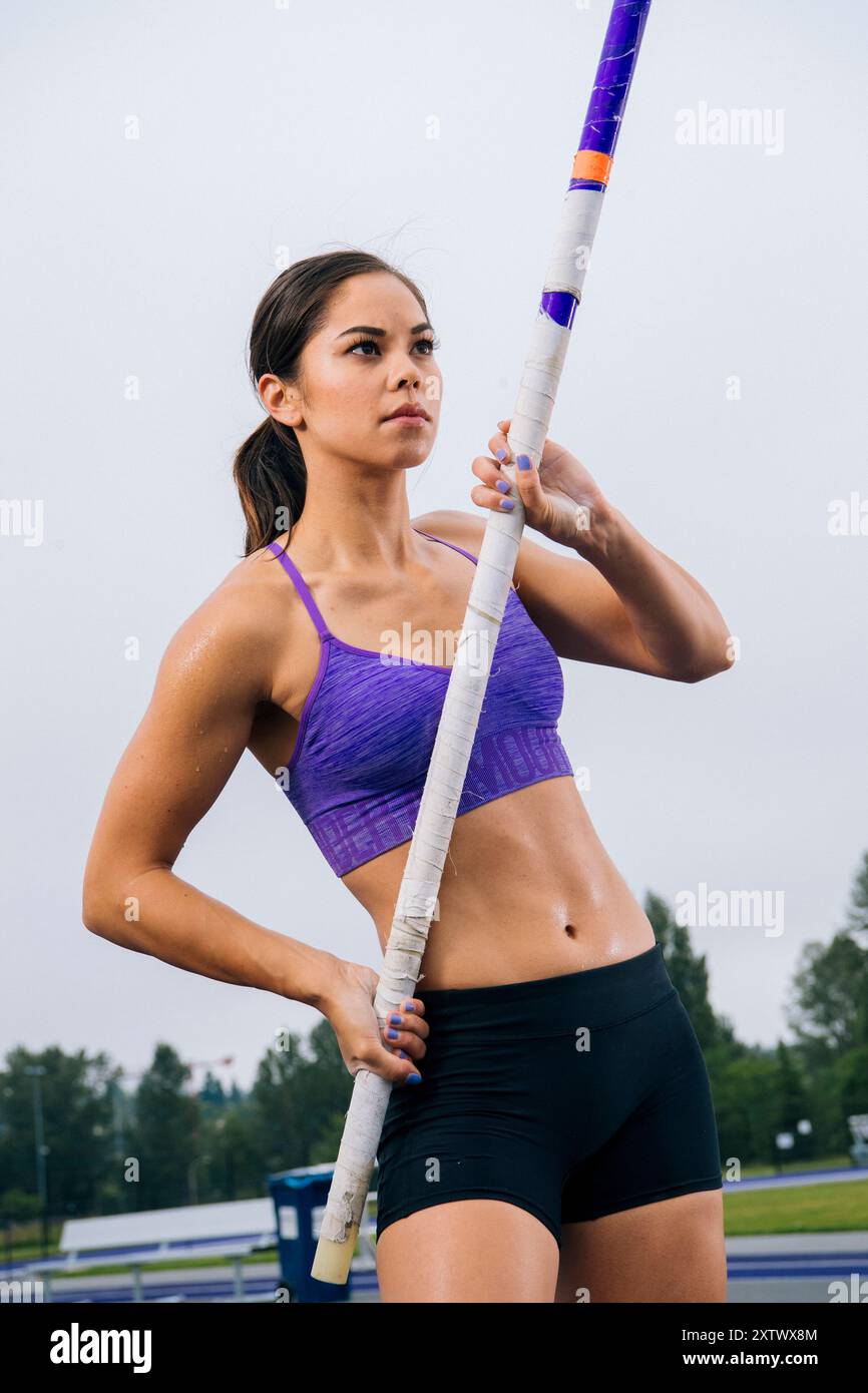 Selbstbewusste Athletin, die einen Stabhochsprung auf einem Rennfeld unter bewölktem Himmel hält. Stockfoto