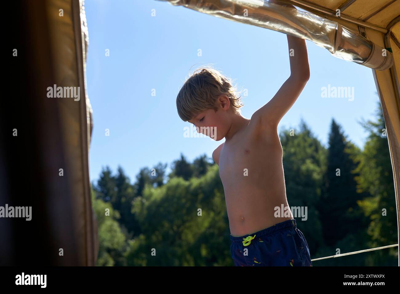 Ein kleiner Junge in Badeshorts blickt von einem schattigen Bereich in eine helle, sonnendurchflutete Landschaft mit Bäumen. Stockfoto