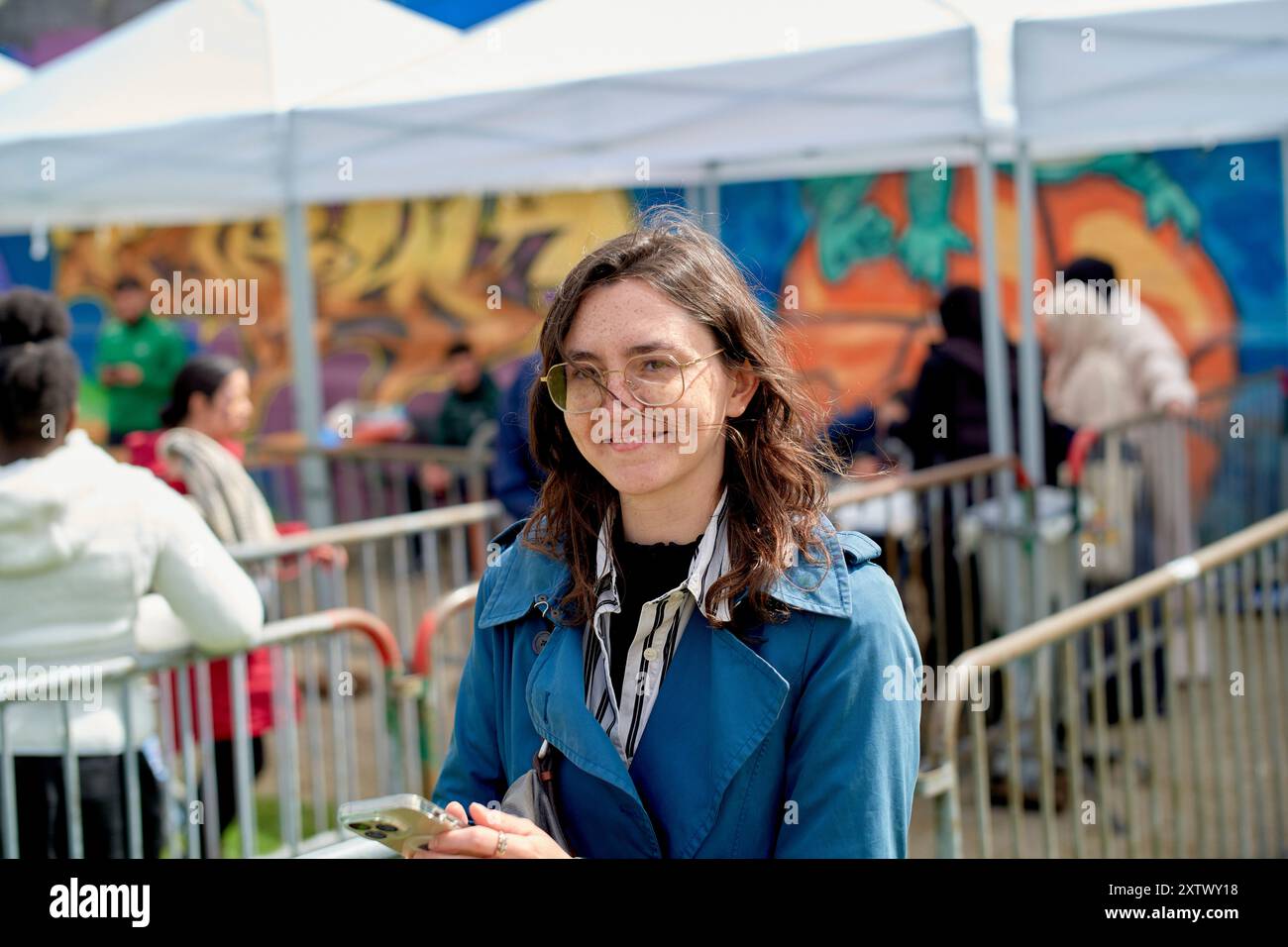 Lächelnde Frau mit Brille, die vor weißen Zelten bei einer Veranstaltung im Freien mit farbenfrohen Wandgemälden im Hintergrund steht. Stockfoto