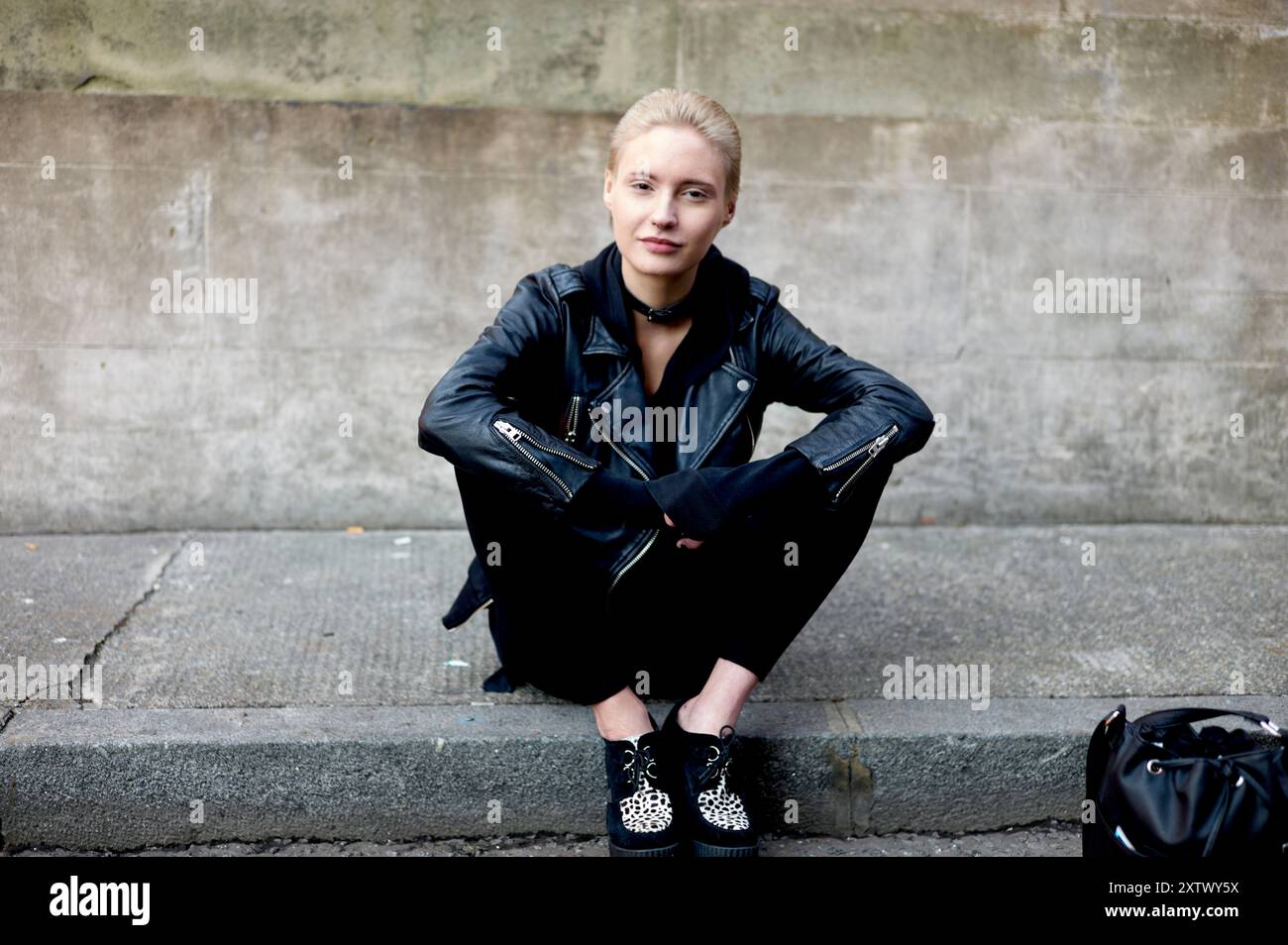 Eine junge Frau in einer schwarzen Lederjacke, die auf einem Bordstein sitzt, ihre Beine überkreuzt und einen selbstbewussten Look hat. Stockfoto