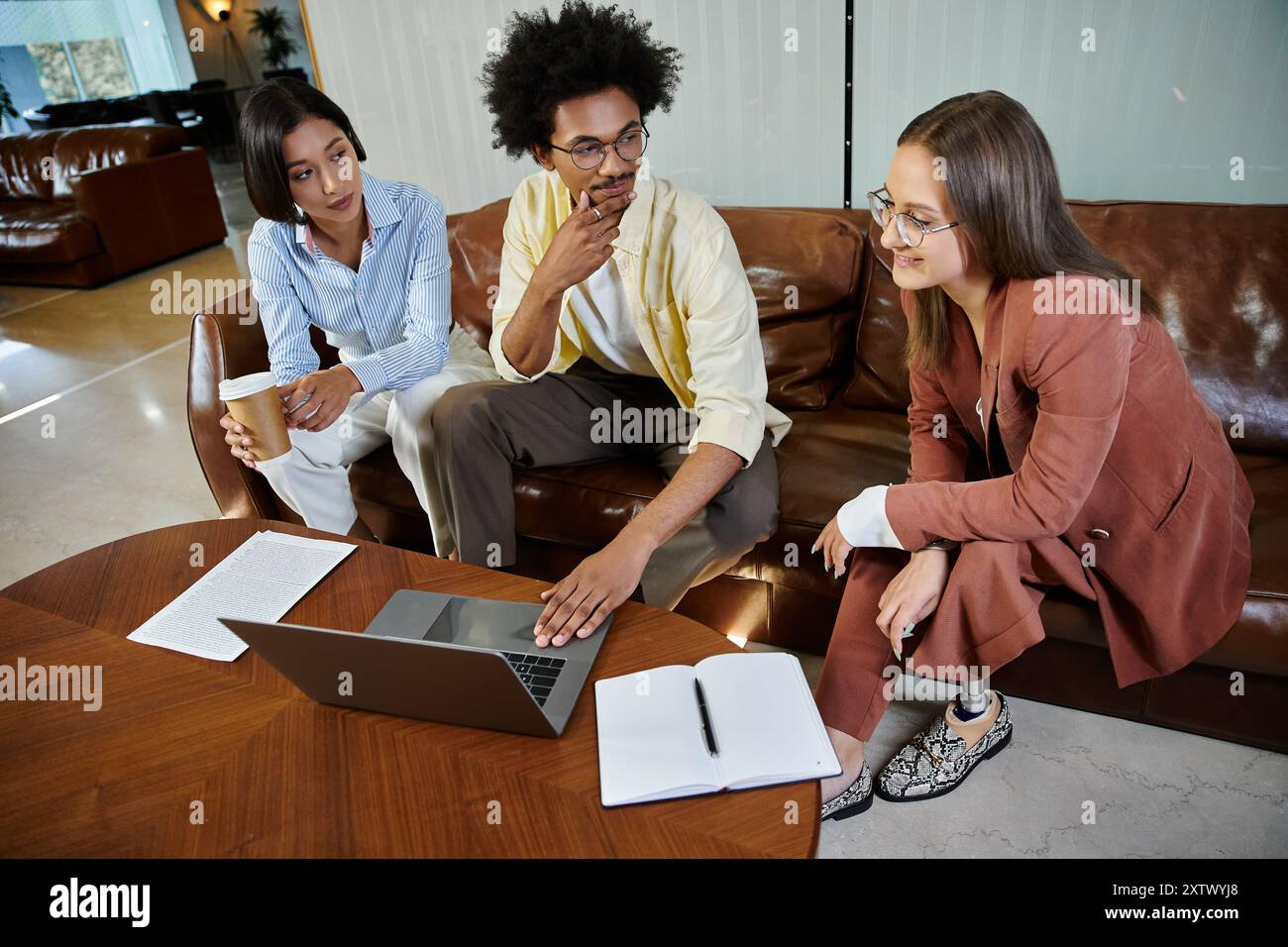 Drei Kollegen treffen sich in einer modernen Bürolobby und diskutieren Ideen, während sie ein Notebook und ein Notebook verwenden. Stockfoto