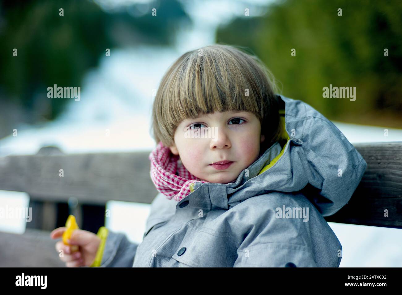 Kleines Kind, das draußen auf einer Bank sitzt, einen grauen Mantel mit einem farbenfrohen Schal trägt und einen gelben Gegenstand hält. Stockfoto