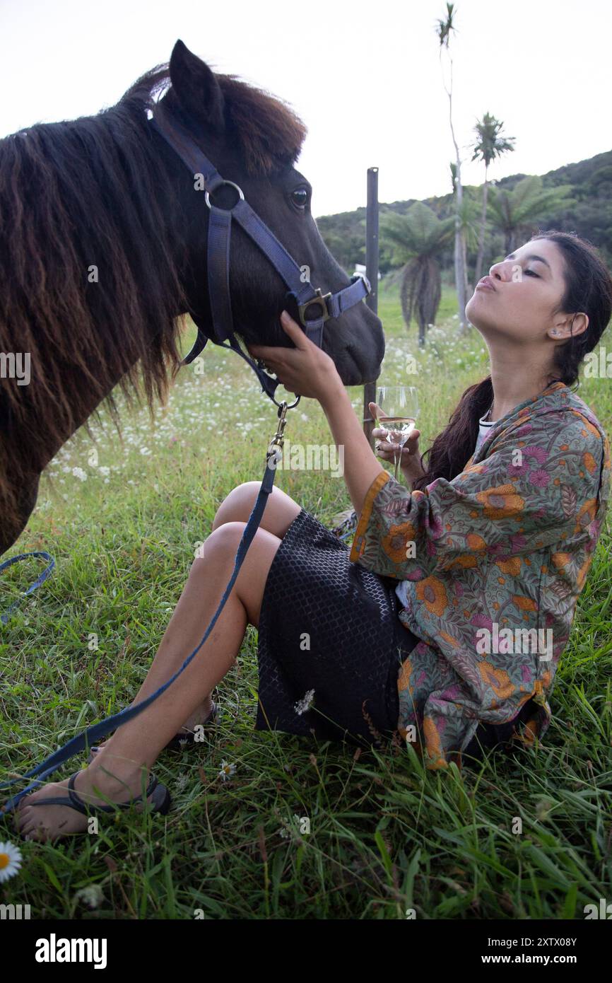 Eine Frau in einem Blumenhemd und einem schwarzen Rock genießt einen Moment mit einem braunen Pferd in einem üppigen grünen Feld, Neuseeland Stockfoto