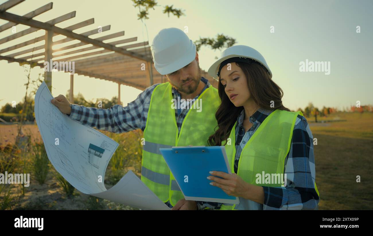 Kaukasische Bauunternehmer Ingenieure Kollegen weibliche Ingenieurinspektorin schreiben Bericht Hardhat männlicher Arbeiter Investor zeigen Bauplan Stockfoto
