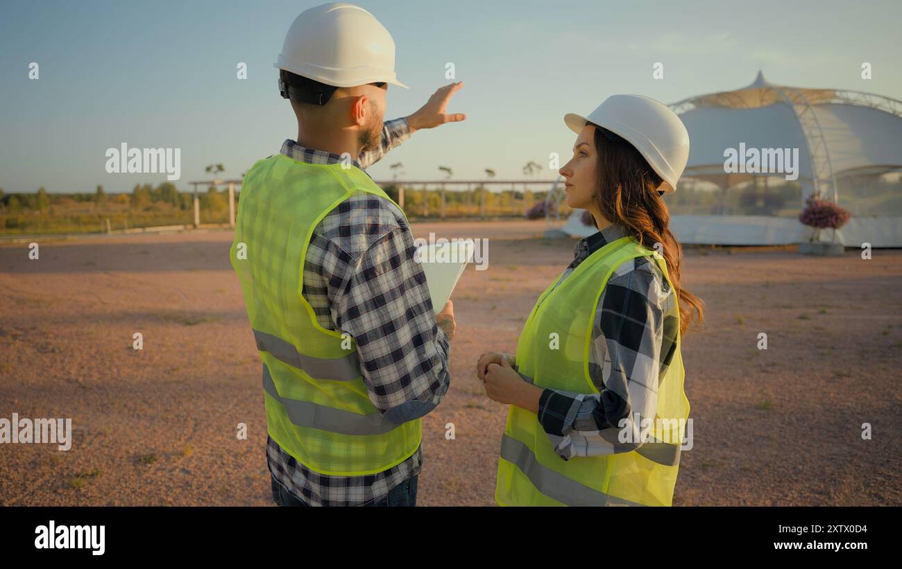 Ein paar kaukasische Techniker, die Arbeitskleidung sprechen. Mann Inspektor Hold Checkliste Show Plan Gesten Frau Ökologie Ingenieur diskutiert Talk Building Stockfoto