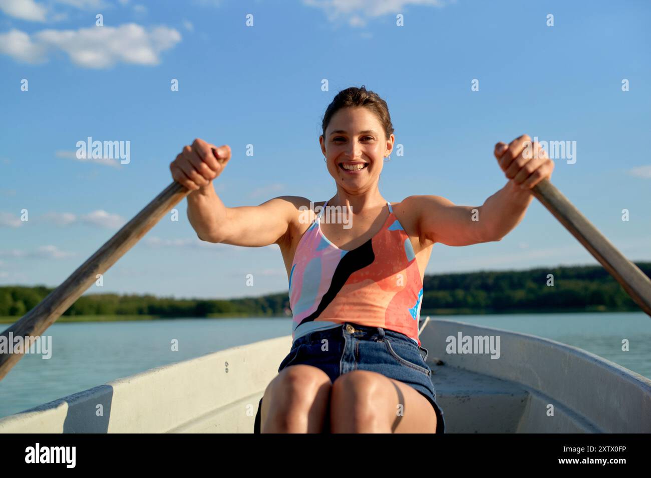 Lächelnde Frau rudert ein Boot an einem sonnigen Tag mit klarem blauen Himmel und einem See im Hintergrund. Stockfoto