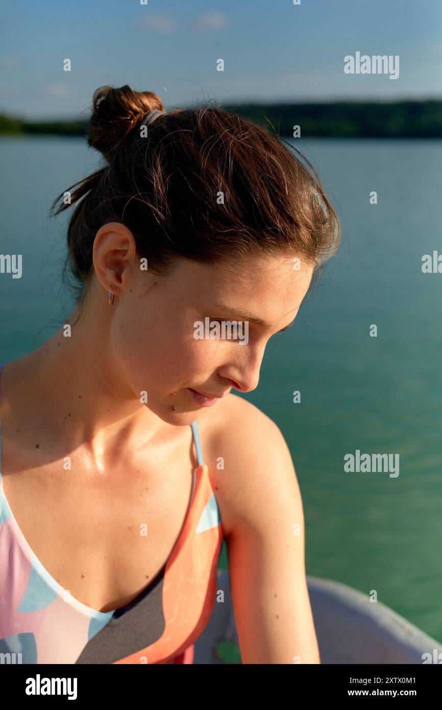 Eine junge Frau mit ihren Haaren in einem Brötchen, die einen Badeanzug trägt, schaut nachdenklich nach unten vor einem Hintergrund von ruhigem Wasser und einer fernen Küste. Stockfoto