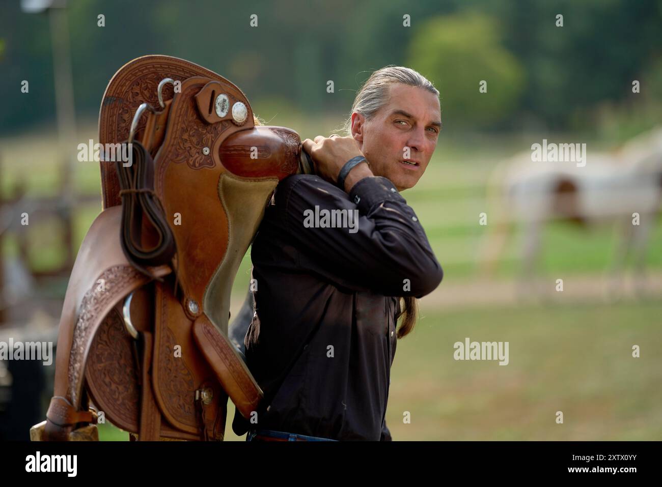 Ein Mann mit silbernen Haaren lehnt sich auf einen Sattel und ruht auf einem Zaun bei einer Reitveranstaltung. Stockfoto