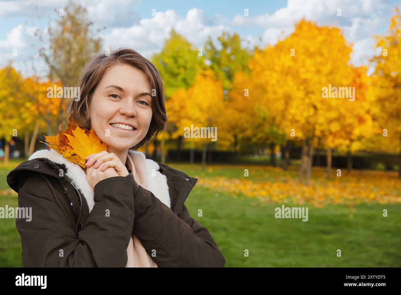 Lächelnde Frau in einer Jacke hält Herbstblätter nahe ihrem Gesicht, stehend in einem Park mit leuchtend gelben Bäumen im Hintergrund Stockfoto