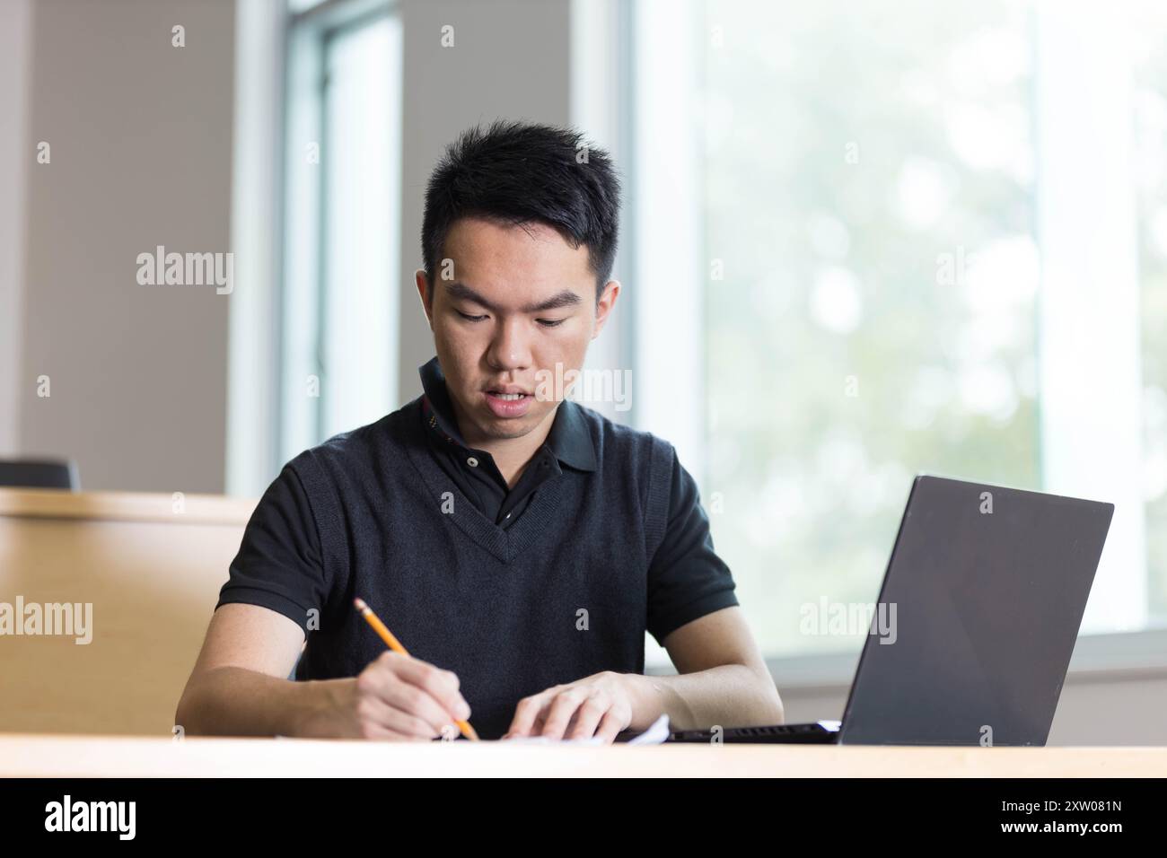 Ein junger Student sitzt gerade an einem Tisch und schreibt mit seinem Bleistift auf ein Blatt Papier mit seinem Laptop neben ihm Stockfoto