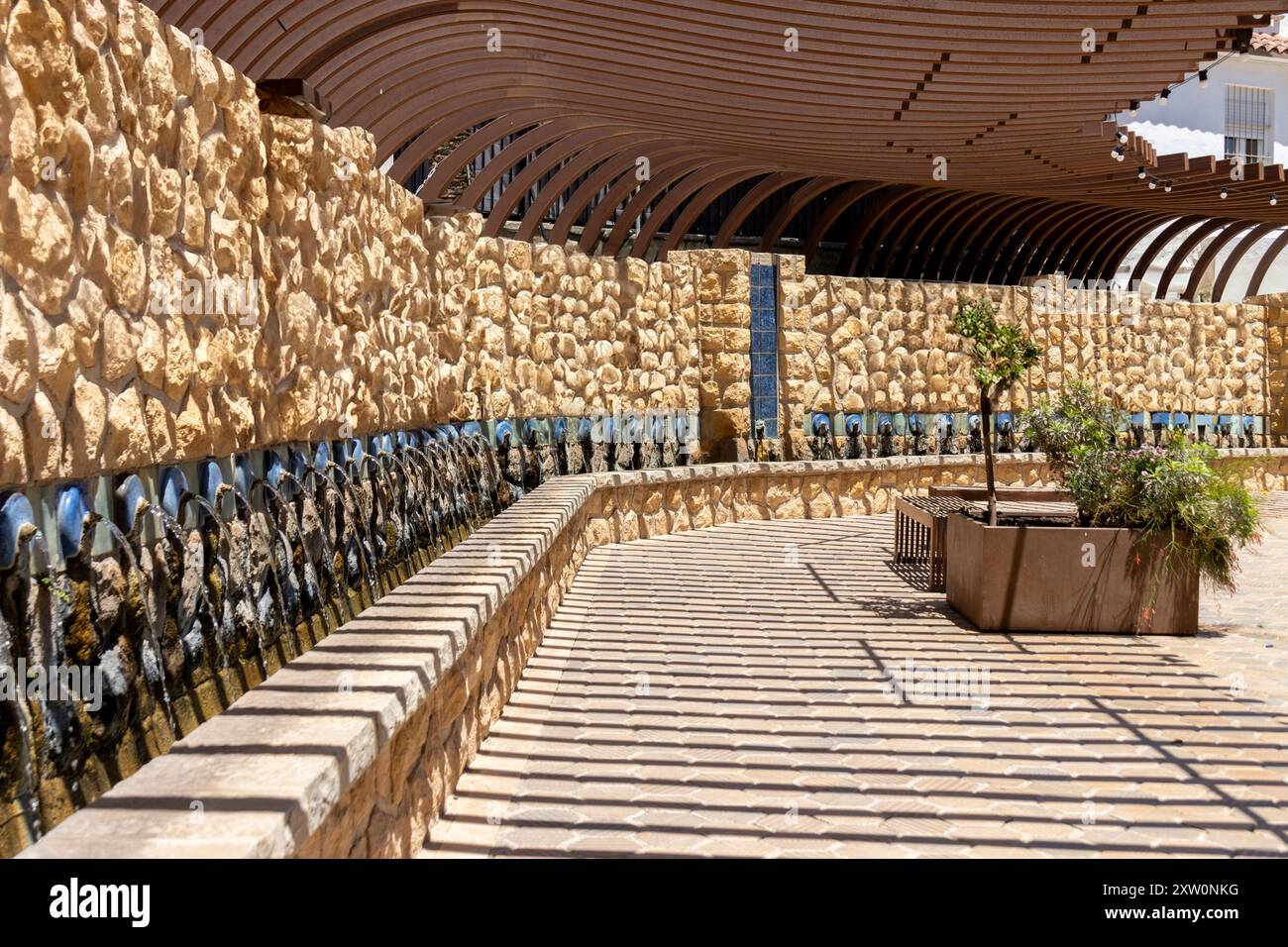 Ein langer Brunnen mit Steinmauern unter einer bogenförmigen Holzpergola in einem öffentlichen Außenbereich mit viel Sonnenlicht. Stockfoto