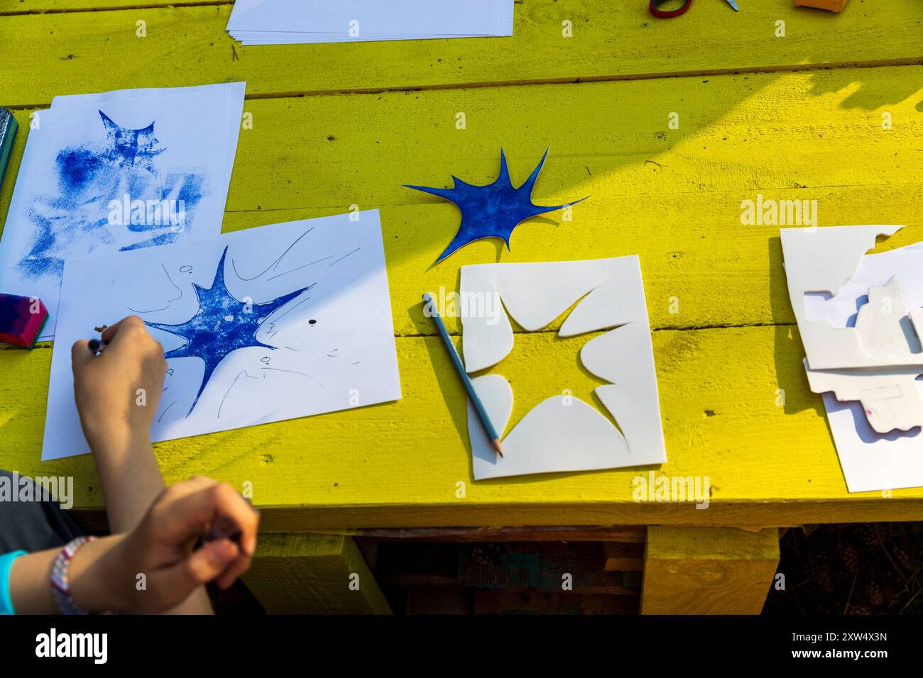 Zeichnungen und Schablonen liegen auf einem gelben Holztisch. Die Hände einer Frau halten einen Bleistift. Stockfoto