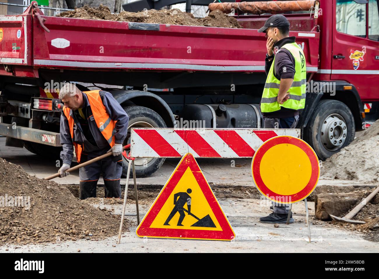 en bei der Arbeit, in Schutzkleidung gekleidet, schwere Maschinen bedienen, während ein Arbeiter einen Graben aushebt, um Sicherheit und Teamarbeit auf der Baustelle zu gewährleisten Stockfoto