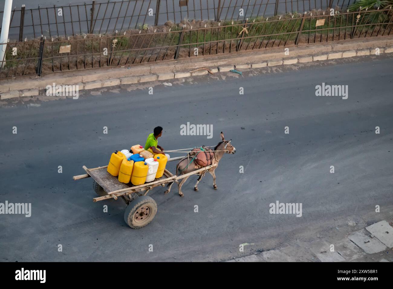 Im ländlichen Afrika betreibt ein junger Mann ein Geschäft, das Wasser in bunten Kanistern verteilt, mit einem primitiven Holzanhänger, der von einem Esel gezogen wird Stockfoto