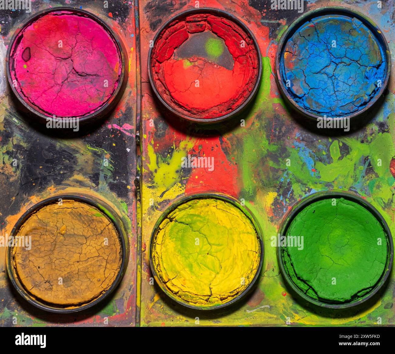 Ein Nahbild einer gut verwendeten Aquarellpalette mit sechs kreisförmigen Wells, die mit getrockneter und gerissener Farbe gefüllt sind. Die Farben sind in lebendigen Tönen gehalten Stockfoto
